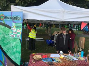 A stall-holder educating at a stand at Envirolution festival while a volunteer wearing a high-vis fixes the stand.